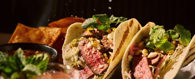 lime being squeezed over 2 meat tacos, next to a bright red cocktail