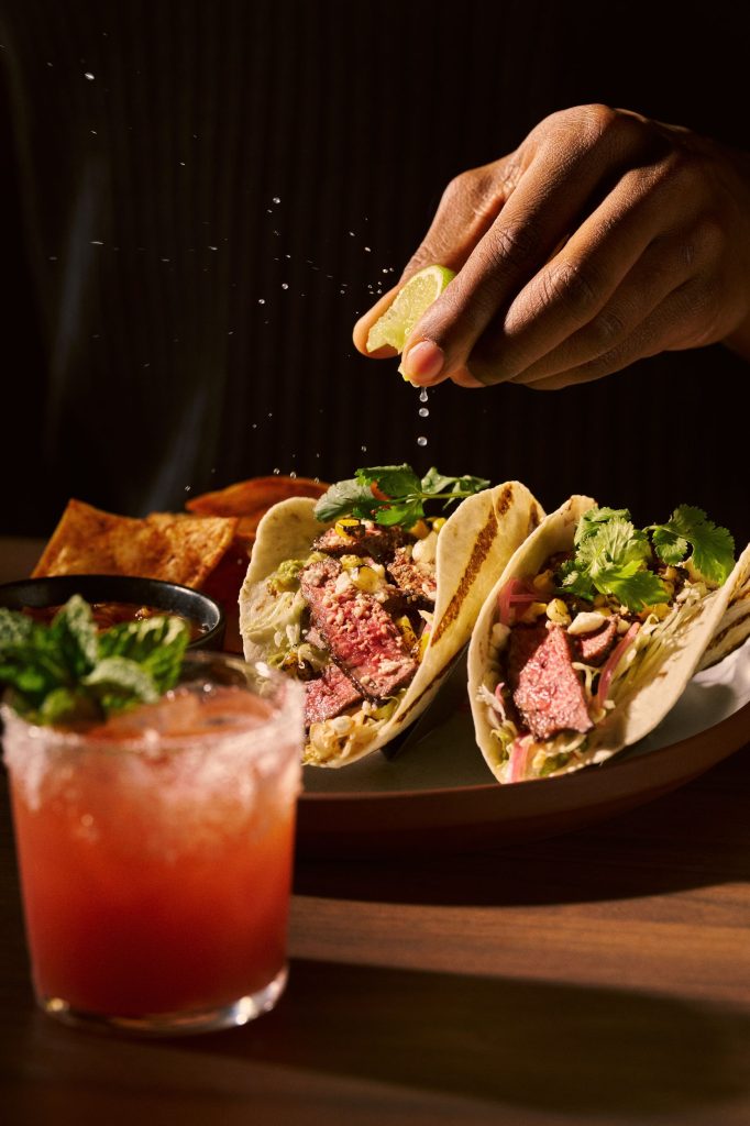 lime being squeezed over 2 meat tacos, next to a bright red cocktail