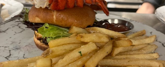 Burger topped with a lobster tail skewer served on a plate with french fries