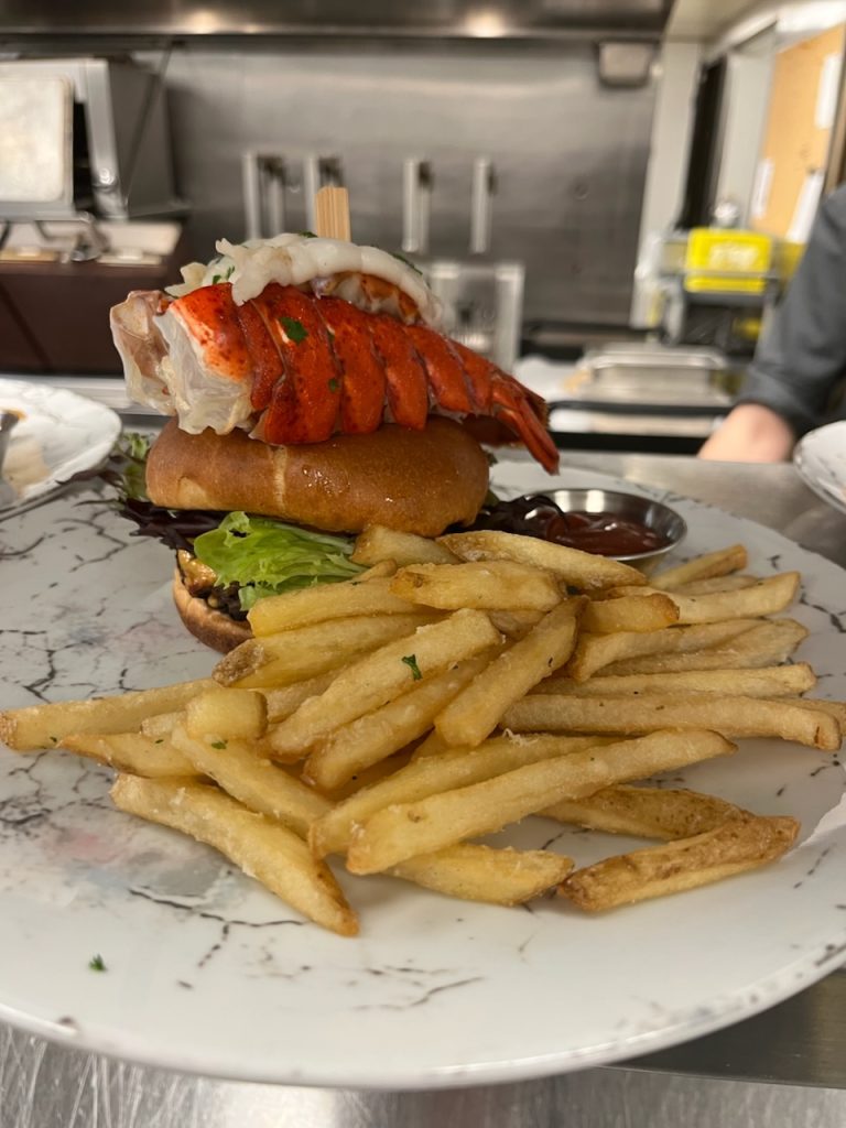 Burger topped with a lobster tail skewer served on a plate with french fries