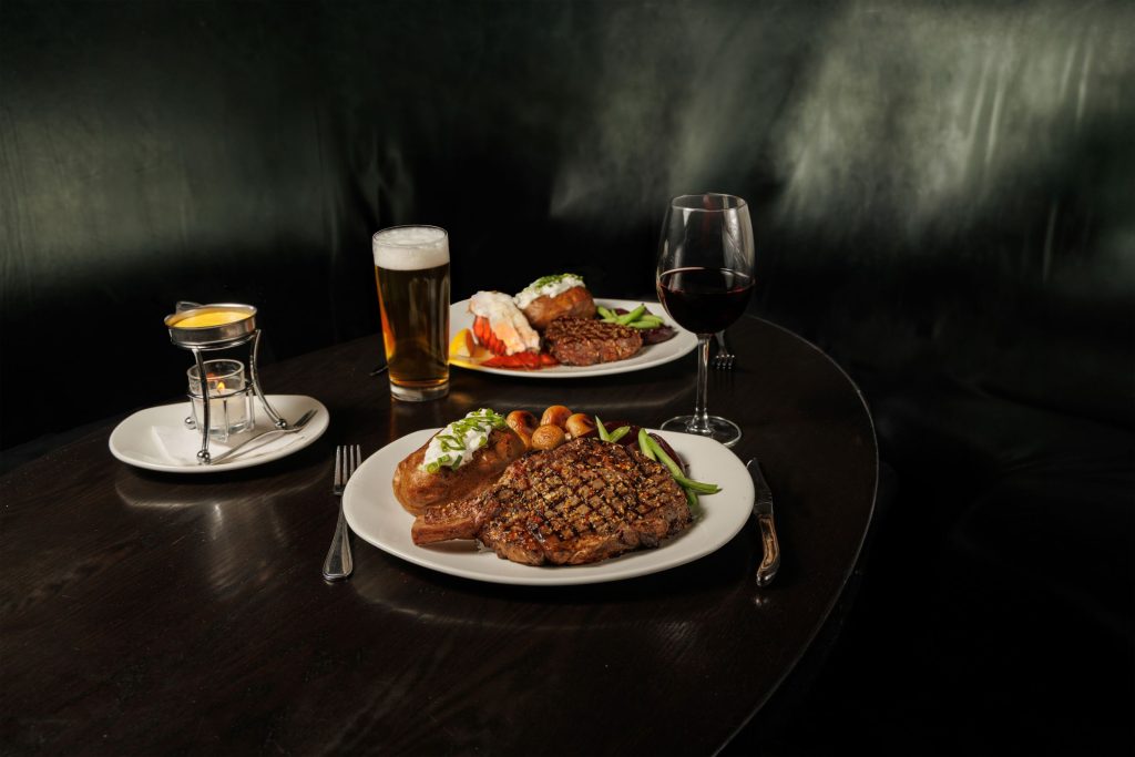 Nicely seared steak on a white plate beside a glass of red wine and a glass of beer. In the background another dish with baked potato and steak.