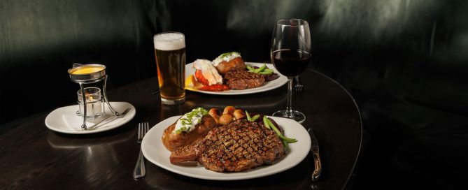 Nicely seared steak on a white plate beside a glass of red wine and a glass of beer. In the background another dish with baked potato and steak.