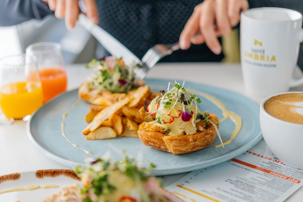 OEB's Truffled Squash Benny on a light blue plate, with a knife and fork poised to cut into it. Beside the plate are two glasses of juice