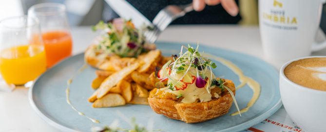 OEB's Truffled Squash Benny on a light blue plate, with a knife and fork poised to cut into it. Beside the plate are two glasses of juice